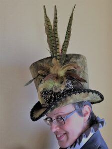 Profile headshot of Lisa Hager, a white woman with short brown hair and glasses on, wearing a green velveteen hat with very tall feathers