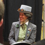 A white person with short, brown hair looks to the side while dressed in a gray coat and hat with a green waistcoat and cravat